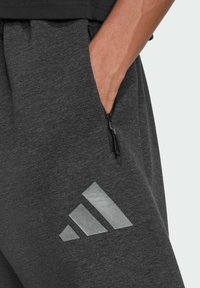 Tmavo šedé teplákové nohavice so zipsovým vreckom a veľkým, odrážajúcim, trojuholníkovým logom vpredu. Mäkká látka s voľným strihom.