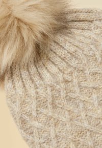 Beige knitted hat featuring a large, fluffy faux fur pom-pom. The texture is soft with a cable knit pattern throughout.