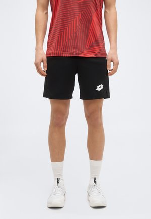 Man wearing black athletic shorts with white logo, red patterned shirt, white crew socks, and white athletic shoes, standing on plain background.