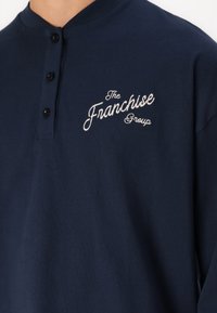 Tamsiai mėlynas medvilninis henley marškinėliai su apvalia iškirpte, trimis juodais mygtukais ir balta siuvinėta užrašu: "The Franchise Group."