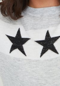 Grauer Pullover mit rundem Ausschnitt, der zwei schwarze Stern-Applikationen auf der Brust hat. Weiche Textur mit einem dezenten Strickmuster.