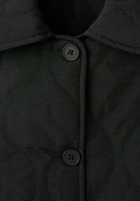 Material negru matlasat cu modele de cusaturi curbate, având două butoane mari negre și un guler lat pe o piesă de îmbrăcăminte.