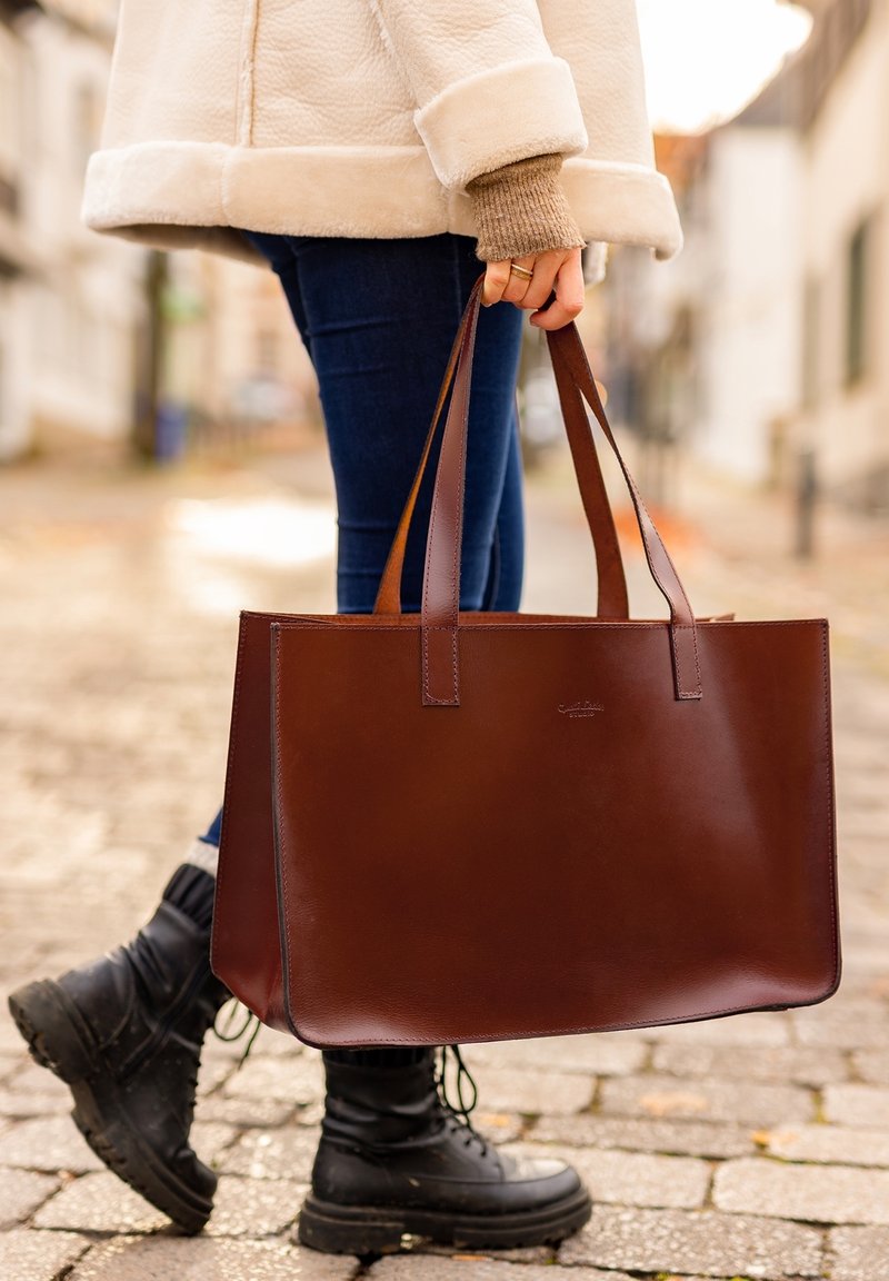 Personne portant une veste beige, un jean bleu et des bottes noires tenant un grand sac cabas en cuir marron dans une rue pavée.