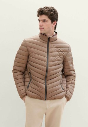 Veste rembourrée marron avec un col montant, design matelassé horizontal et accents de fermeture éclair noirs. Portée sur un pantalon beige.