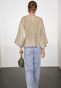 Blouse en tissu beige froissé à manches larges, associée à un jean en denim bleu clair. Le mannequin tient un petit sac vert.