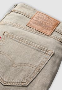 Jeans de mezclilla beige con una superficie texturizada, que cuentan con un parche de cuero marrón y un distintivo bordado en el bolsillo trasero.