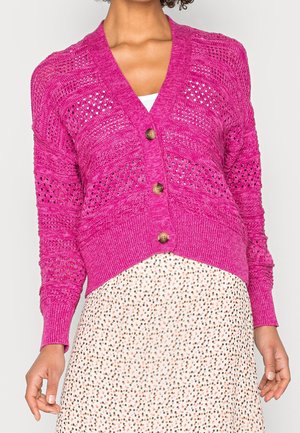 Kvinde iført lys pink strikket cardigan med tre store knapper over hvid top og hvid nederdel med prikker, hænderne afslappet ned langs siden.