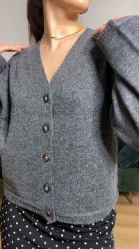 Siv pleteni kardigan z globokim V-izrezom, s petimi okroglimi gumbi in rebrastimi robovi, kombiniran s črno krilo z belimi pikami.