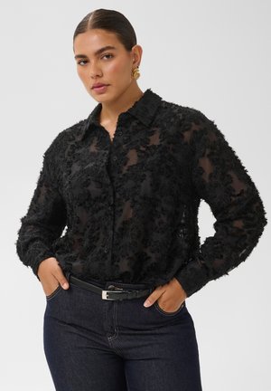 Femme aux cheveux tirés en arrière, portant des boucles d'oreilles en or, un chemisier noir transparent texturé et un jean foncé avec une ceinture noire, mains dans les poches.