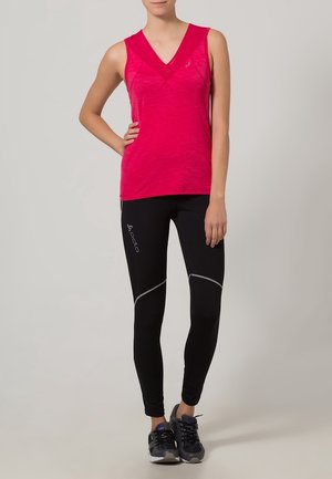 Kobieta w jasnoróżowym bezrękawniku z dekoltem w serek, czarnych dopasowanych legginsach z logo i odblaskowymi detalami oraz czarnych butach sportowych.