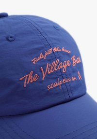 Modrá čepice vyrobená z lehkého materiálu, na přední straně s vyšitým textem v růžové barvě: "Cítí se jako doma - The Village Bar - scalpers co."