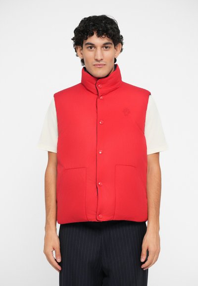 Red insulated vest with a high collar, front snap buttons, and two large pockets. Features a subtle logo on the chest, worn over a light shirt.