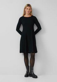 Robe noire en tricot côtelé avec des manches longues, une jupe évasée et un col rond. Associée à des bottines noires et des collants transparents.