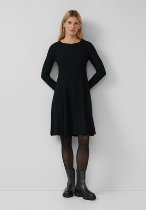 Robe noire en tricot côtelé avec des manches longues, une jupe évasée et un col rond. Associée à des bottines noires et des collants transparents.