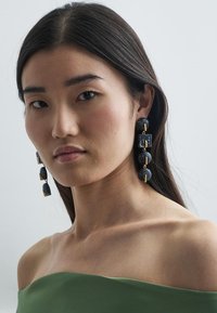 Orecchini in pietra nera con forme circolari e rettangolari, collegati da accessori in tonalità oro, indossati da una modella con lunghi capelli scuri.