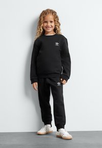 Fekete póló és melegítő nadrág szett, pamutból készült, fehér Adidas logóval. A gyermek fehér sportcipőt visel, gumis talppal. Egyszerű háttér.