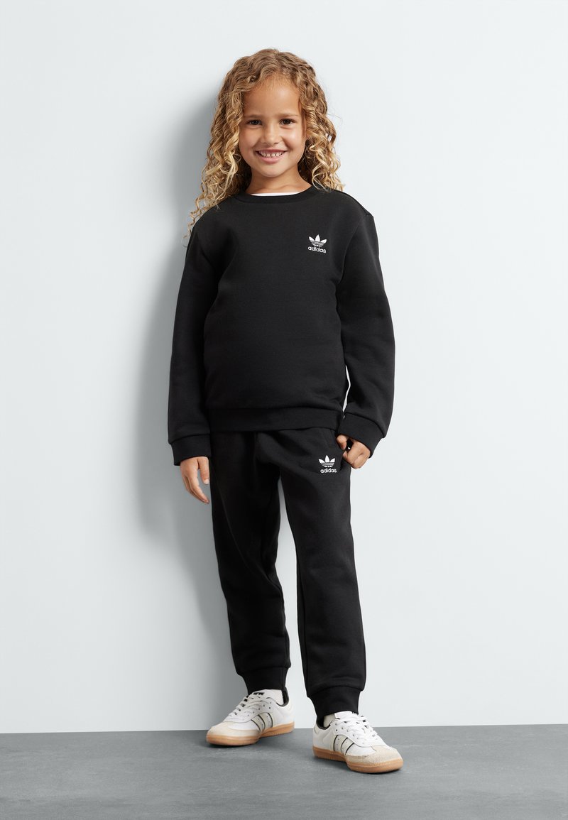 Fekete póló és melegítő nadrág szett, pamutból készült, fehér Adidas logóval. A gyermek fehér sportcipőt visel, gumis talppal. Egyszerű háttér.