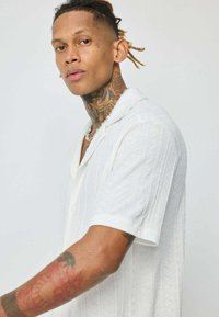 Chemise blanche à manches courtes avec un motif texturé, présentant un col en pointe et un patte de boutonnage. Un modèle tatoué se tient contre un fond clair.