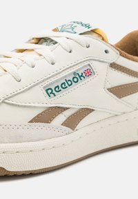 Fehér bőr sneaker bézs részletekkel, texturált gumitalppal, klasszikus fűzős záródással és zöld feliratos Reebok logóval.