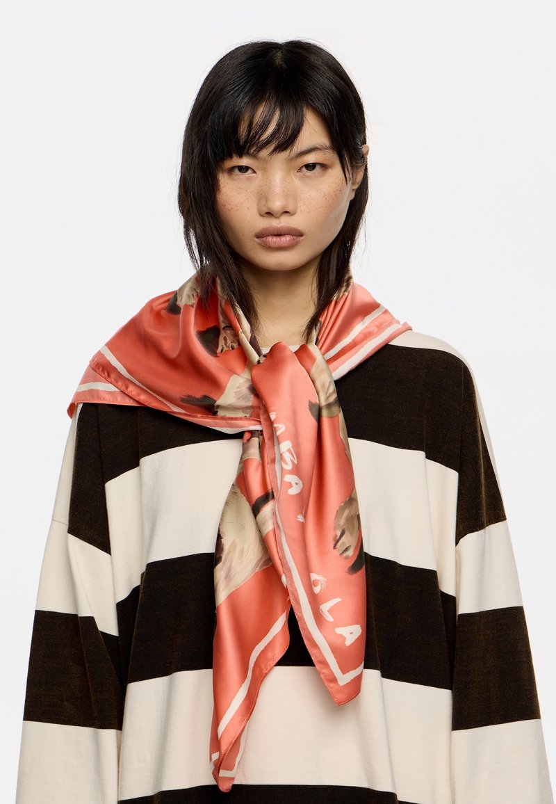 Foulard di seta in corallo, con stampa di ritratto e testo bianco, annodato attorno al collo. Indossato sopra una maglia a righe bianco e nero.