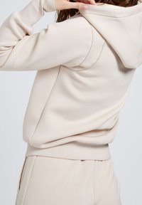 Sudadera con capucha beige de textura suave, con costuras detalladas a lo largo del hombro y un corte relajado.