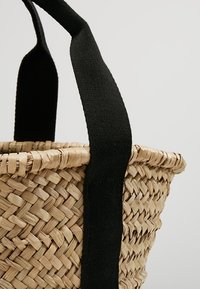 Cesta di paglia intrecciata con ampi manici in tessuto nero attaccati al bordo, mostrata su uno sfondo chiaro e semplice.