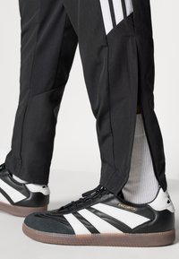 Μαύρο αθλητικό παντελόνι με λευκές ρίγες και φερμουάρ στους αστραγάλους, συνδυασμένο με ασπρόμαυρα παπούτσια ποδοσφαίρου με υφασμένο πάνω μέρος και καφέ σόλα.