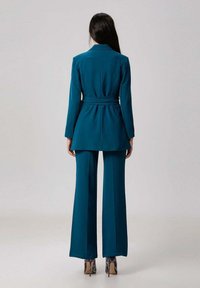 Femme aux longs cheveux noirs portant un blazer ceinturé teal et un pantalon large assorti, se tenant de dos, sur un fond clair et uni.