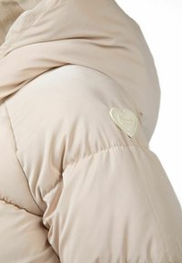 Lyse beige pufferjakke har en glat, skinnende overflade med et hjerteformet logo på ærmet. Designet inkluderer en hætte og sammensnørede manchetter.