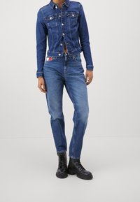 Modrá džínová bunda s límečkem, dvěma náprsními kapsami, v kombinaci s modrými džínovými kalhotami s vysokým pasem a černými šněrovacími kotníkovými botami.