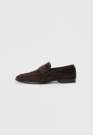 Mocasines de ante marrón con una textura suave, que presentan un ribete cosido y un acento de tiras, montados sobre una suela baja negra.