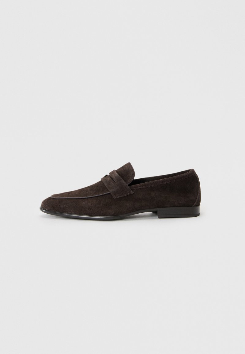 Mocasines de ante marrón con una textura suave, que presentan un ribete cosido y un acento de tiras, montados sobre una suela baja negra.