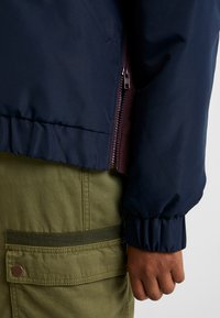 Chaqueta azul marino con cierre de cremallera y puños fruncidos, combinada con pantalones cargo verde oliva que presentan un detalle de bolsillo con parche.