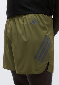 Ελαφριά αθλητικά σορτς σε ελιά από ύφασμα ελαφριάς σύνθεσης, με γκρι διακοσμητικές τρεις ρίγες και πλευρικά ανοίγματα για μεγαλύτερη ευκινησία.