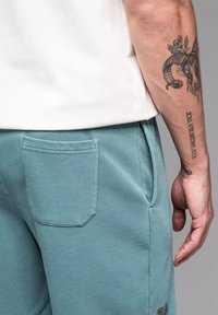 Shorts en coton teal avec une poche arrière et une poche d'entrée latérale, portés avec une chemise claire à manches courtes. Tissu texturé et lignes épurées.