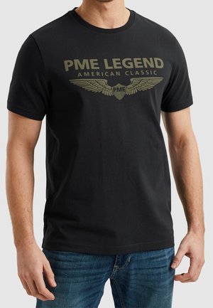 Mand iført en sort t-shirt med "PME LEGEND AMERICAN CLASSIC" og vingeemblem trykt i olivengrøn, kombineret med blå jeans.