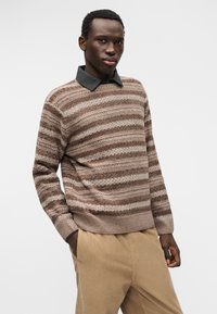 Hnědý pruhovaný pletený svetr s kulatým výstřihem a žebrovaným lemem, vrstvený přes šedou košili s límečkem. Kapsy jsou viditelné na béžových manšestrových kalhotách.