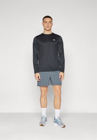 Mans atletiska outfit som består av en långärmad svart tröja och grå shorts. Grå sneakers med blå detaljer, vita strumpor och en enkel design.