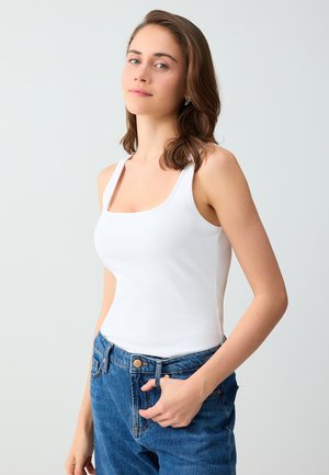 Młoda kobieta o brązowych włosach do ramion, ubrana w biały top i niebieskie dżinsy, stoi na jasnym jednolitym tle.