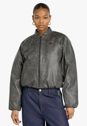 Giovane donna con orecchini a cerchio dorati che indossa una giacca grigio scuro con zip e jeans blu a vita alta con strisce laterali.