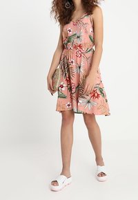 Vestido floral rosa con una cintura ajustada, tirantes finos y una falda acampanada. Combinado con sandalias blancas y un bolso redondo de paja.