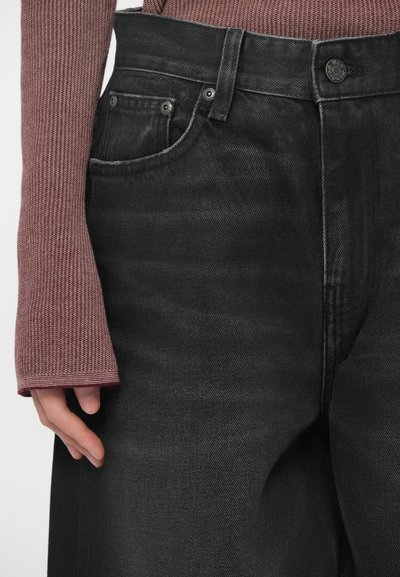 Zajem v neposredni bližini osebe, ki nosi črne hlače z visokim pasom in dolgo sleeve teksturirano roza srajco, z roko sproščeno ob njeni strani.