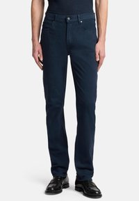 Dunkelblaue Denim-Jeans mit schmaler Passform, Fünf-Taschen-Design, vorderem Knopfverschluss und dezenter Nähedetail. Getragen mit schwarzen Dressschuhen.