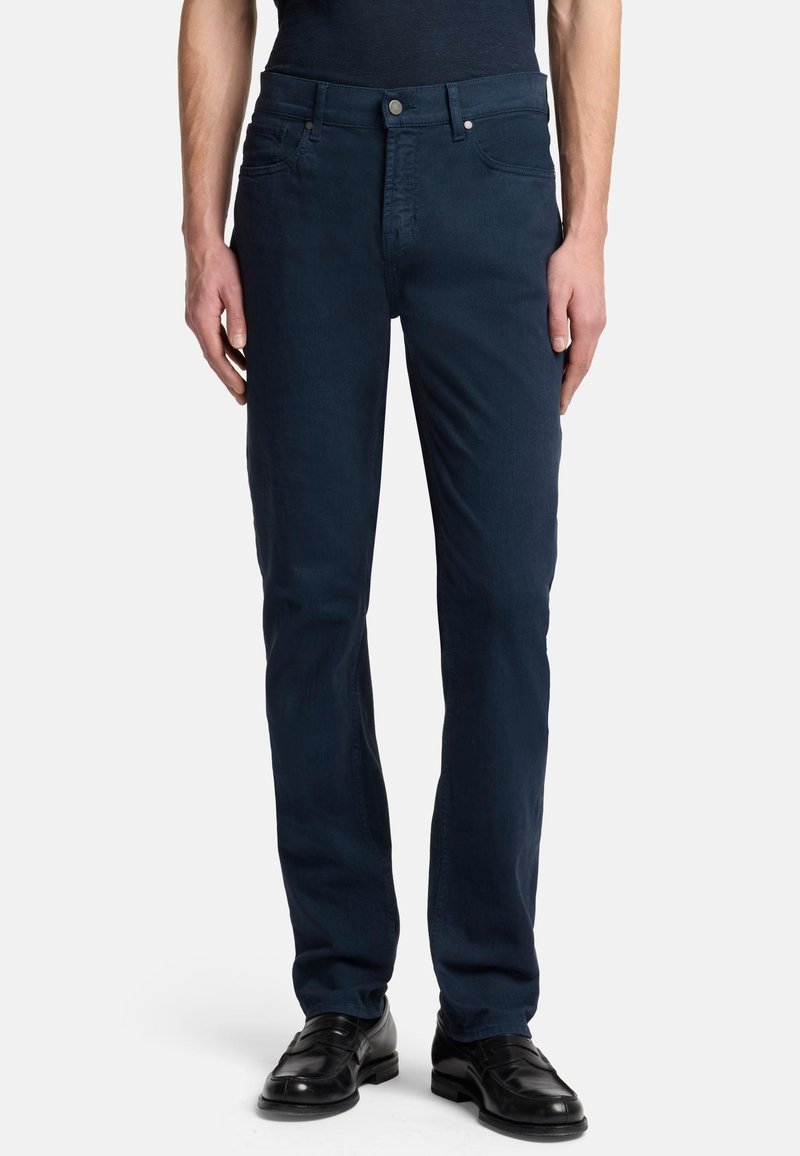 Dunkelblaue Denim-Jeans mit schmaler Passform, Fünf-Taschen-Design, vorderem Knopfverschluss und dezenter Nähedetail. Getragen mit schwarzen Dressschuhen.