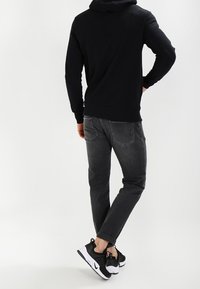 Persona vestida con una sudadera negra, jeans gris oscuro y zapatillas blanco y negras, de pie con una mano en la cadera y las piernas cruzadas.