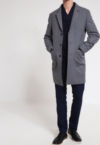 Abrigo de lana gris con solapa English, cierre de botones, bolsillos laterales, llevado sobre un suéter oscuro y pantalones de navidad, combinado con zapatos negros.