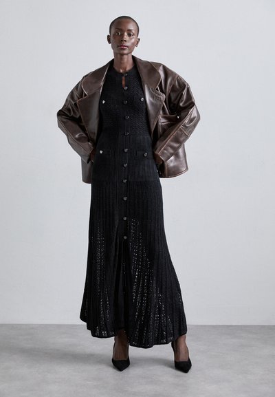 Sort strikket lang kjole med knaplukning foran, ribbet tekstur og to forlommer, lagd under en stor brun læderjakke. Sorte hæle fuldender looket.