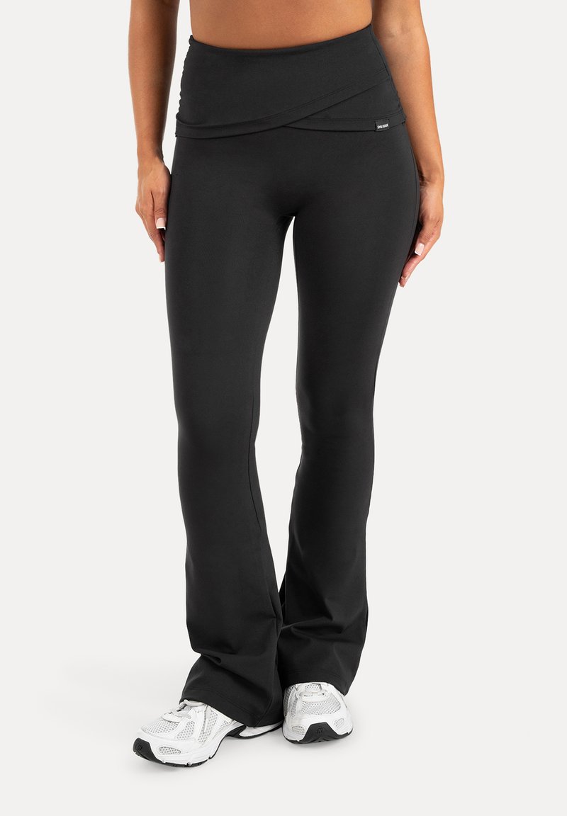 Femme portant un pantalon de yoga noir ajusté avec une taille haute et des chaussures de course blanches, debout devant un fond uni.