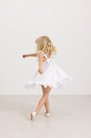Robe blanche avec des bretelles et un ourlet à volants, en tissu léger, jupe évasée, nœud dans le dos très détaillé, associée à des chaussures blanches. Mouvement de rotation.