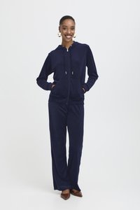 Marineblå velour-träningsdräkt som har en zip-up hoodie med framfickor och matchande vida byxor. Utformad för komfort och rörelsefrihet.